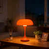 Una lámpara de escritorio naranja con pantalla abombada sobre una mesa de madera en una habitación tenue.
