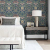 Pared de dormitorio con papel tapiz floral damasco vintage en tonos azules, verdes y dorados, con una lámpara de noche y una 