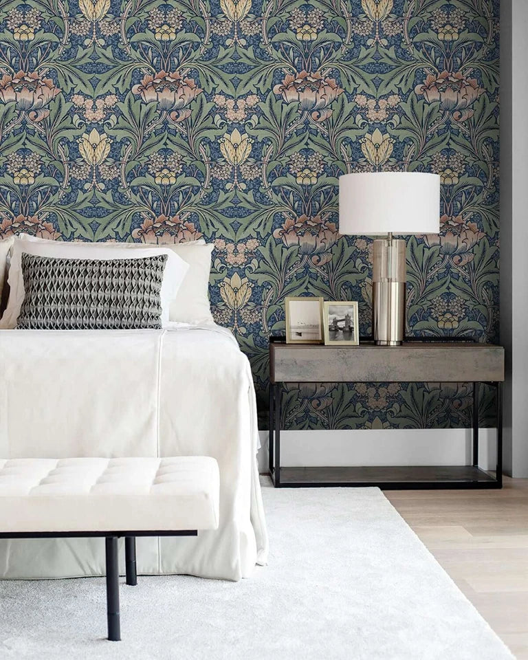 Pared de dormitorio con papel tapiz floral damasco vintage en tonos azules, verdes y dorados, con una lámpara de noche y una 