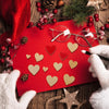 Red felt card with gold heart cutouts and pine needles, surrounded by natural elements and white gloves.