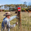 A man in a plaid shirt is using a tool to adjust a red and white solar motion sensor security alarm wire on a wooden post.