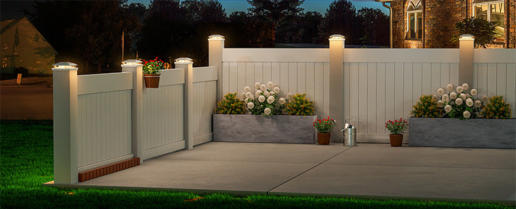 A white wooden fence with illuminated post cap lights and potted flowers in a backyard setting.