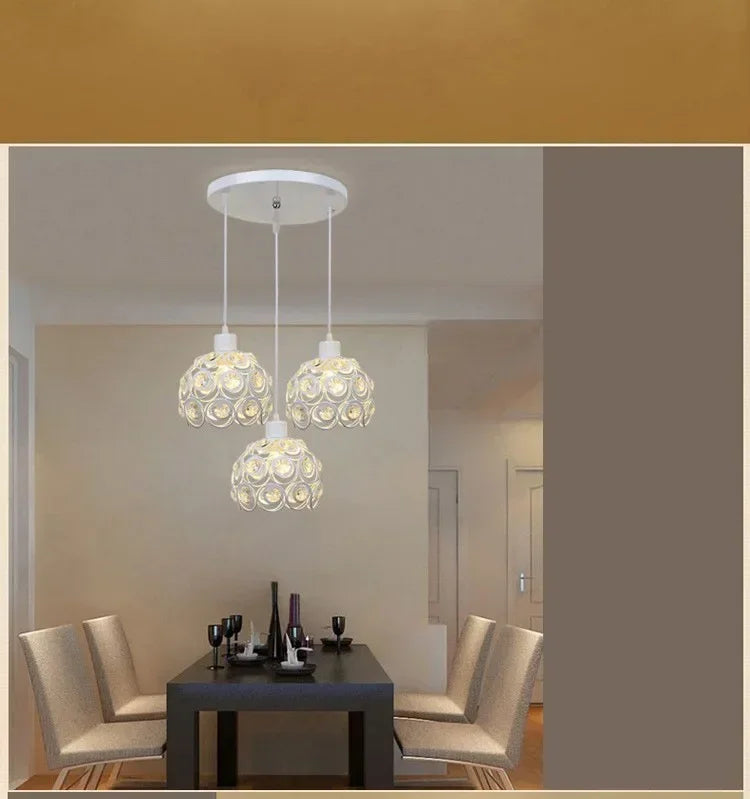 A modern crystal chandelier lamp with three glass globes hangs above a dark table in a well-lit room.