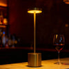 A modern gold desk lamp with a circular LED light illuminates the surface next to a glass of red wine.