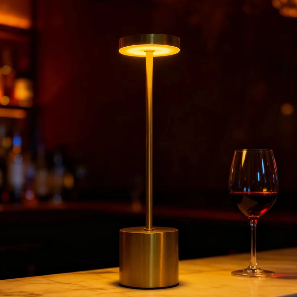 A modern gold desk lamp with a circular LED light illuminates the surface next to a glass of red wine.