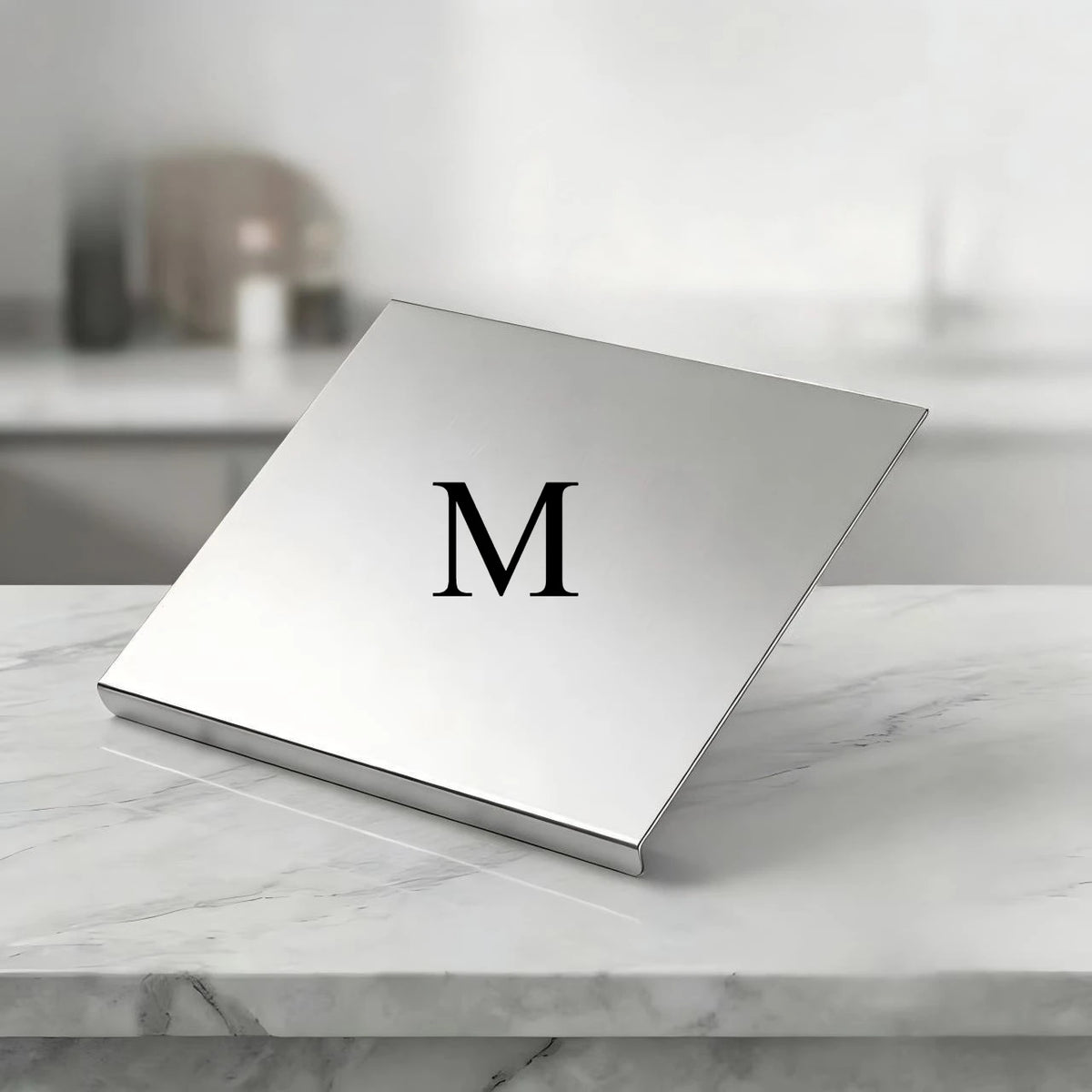 A square stainless steel cutting board with a silver finish and a large black letter M is displayed on a white marble surface