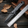 A black multi-functional camping survival knife rests on a wooden surface next to a ruler and autumn leaves.