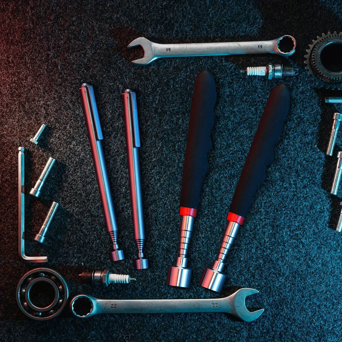 A collection of various metal tools and components including wrenches, drill bits, and magnetic grippers on a dark surface.