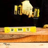 A hand wearing a yellow glove holds a precision magnetic box level next to a wooden surface.