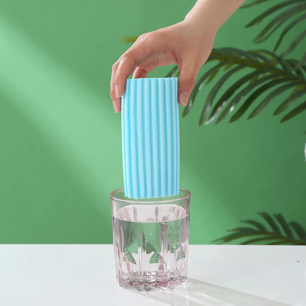 A hand is holding a light blue PVA absorbent cleaning sponge over a glass on a white surface.