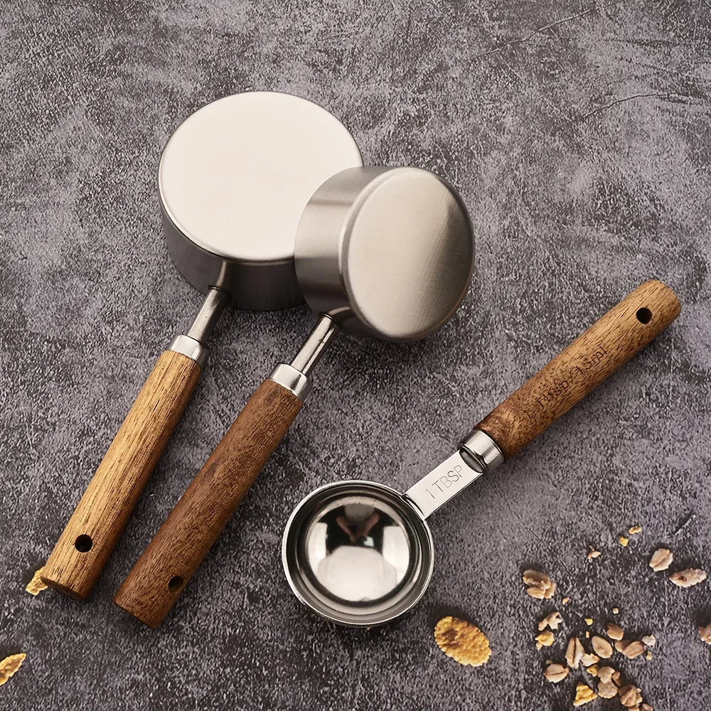 A stainless steel and wooden measuring spoon set with a metal bowl is shown on a gray surface.