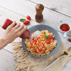 A hand pours red seasoning from a squeeze dispenser bottle onto pasta in a bowl surrounded by fresh tomatoes.