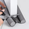A hand is using scissors to cut a section of a gray windproof door draft excluder seal strip.