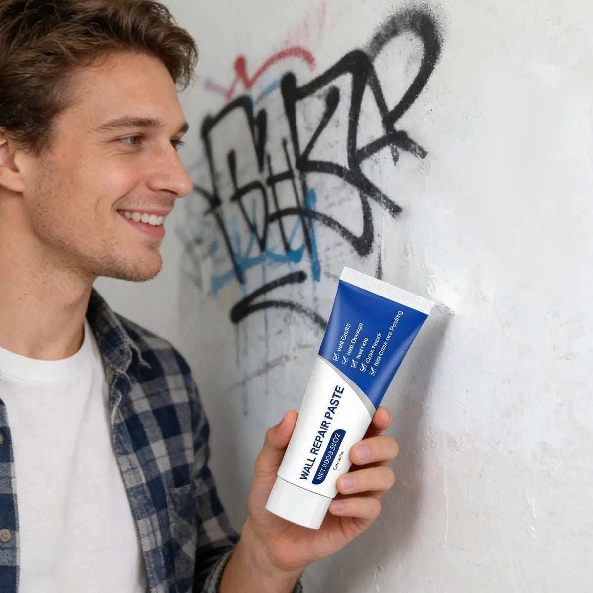 A man holds a tube of Wall Repair Paste against a white wall with black graffiti marks.