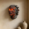 A dark, cracked resin sculpture of a face with glowing orange and yellow internal light mounted on a light-colored wall.