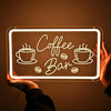 A hand holds a glowing white LED neon sign shaped like 'Coffee Bar' on a dark background.