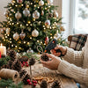 A person uses a hot melt glue gun to apply glue to pine cones on a wooden table in front of a decorated Christmas tree.