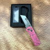 A folding utility knife with a pink handle and stainless steel blade is displayed in a black box on a brown surface.