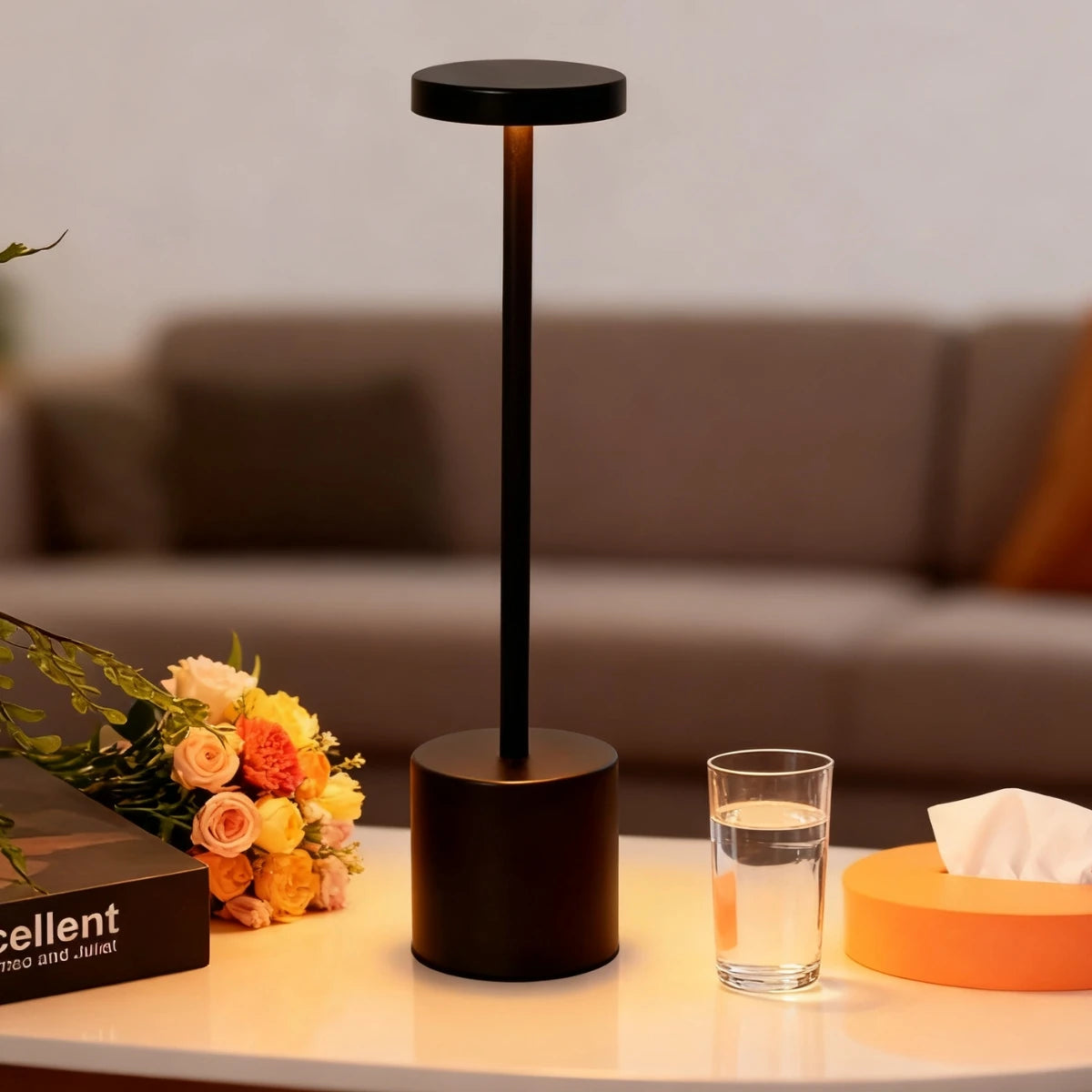 A black desk lamp with a cylindrical base and a round shade stands on a white surface next to a glass of water.
