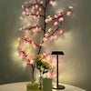 A branch of LED cherry blossom tree lights illuminates pink flowers against a light green wall.