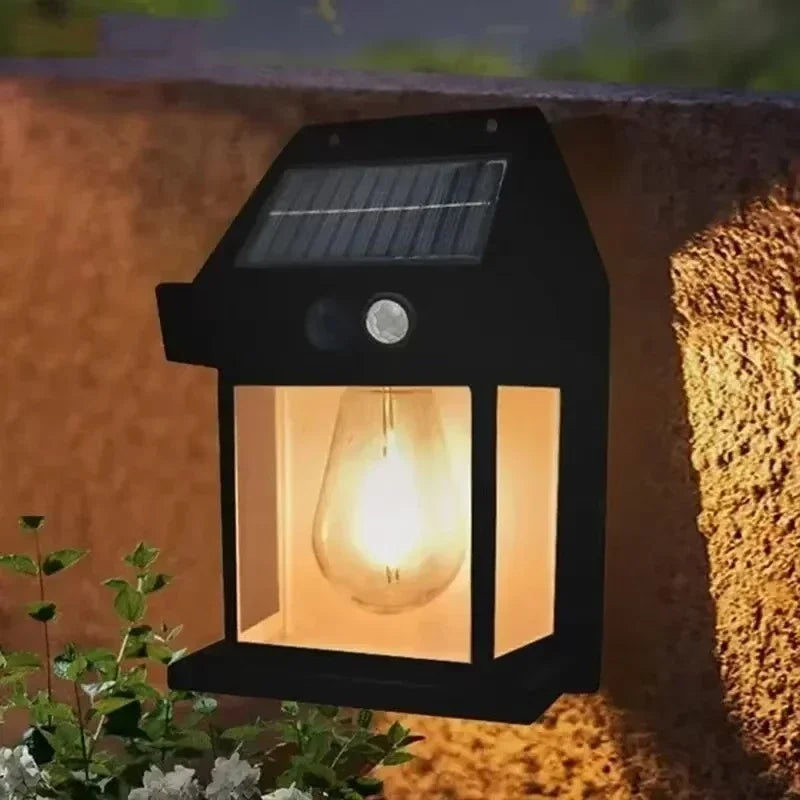 A black solar motion sensor LED wall light with a glowing bulb mounted on an outdoor wall.