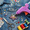 A purple hot melt glue gun and various craft supplies like beads, gems, and scissors are laid on a denim fabric.
