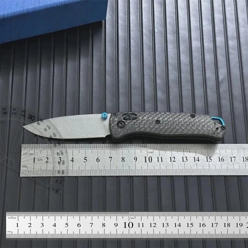 A folding knife with a carbon fiber handle and blade resting on a ruler.