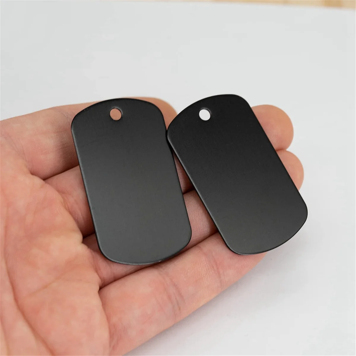 Two black aluminum metal stamping tags are held in a hand against a white background.