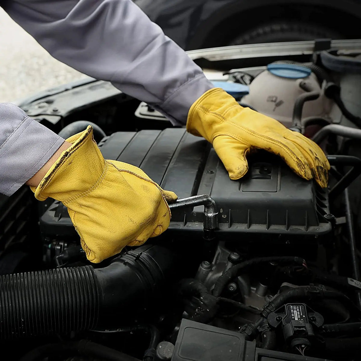 Eine Person mit gelben Kuhfell-Gartenhandschuhen arbeitet im Motorraum eines Fahrzeugs.