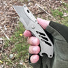 A hand holds a stainless steel folding knife with a serrated blade and textured handle in an outdoor setting.