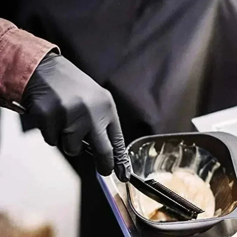 A hand wearing a black nitrile glove is using a black brush to apply a cream or paste inside a dark bowl.