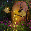A solar fairy watering can light illuminates plants in a garden setting.