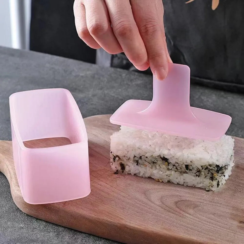 A hand is pressing a pink silicone mold onto a piece of rice, demonstrating a DIY sushi and onigiri making kit.