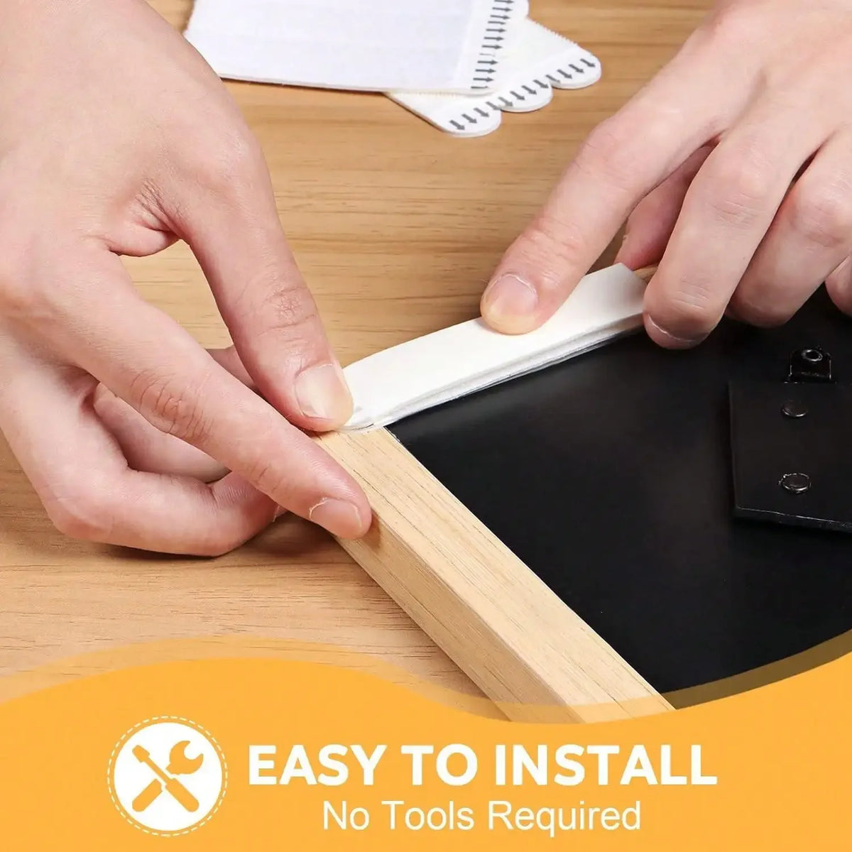 Hands are applying double-sided photo frame adhesive strips to a black frame on a wooden surface.