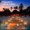 A wooden walkway lined with glowing torch lights leading toward a tropical silhouette at sunset.