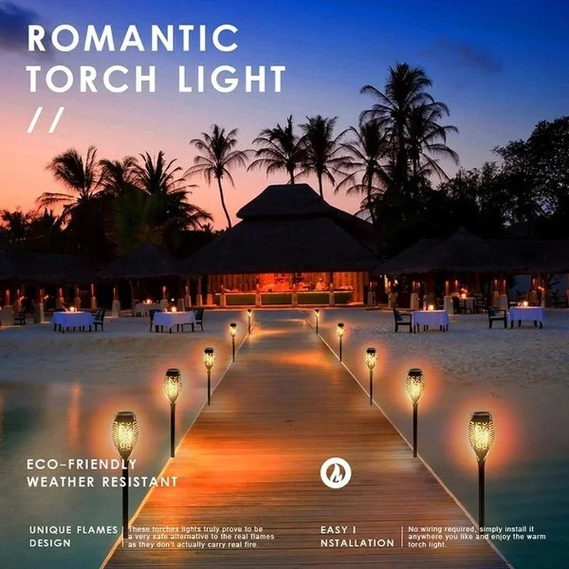 A wooden walkway lined with glowing torch lights leading toward a tropical silhouette at sunset.