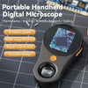 A portable handheld digital microscope with a black body and orange switch, resting on a dark surface.
