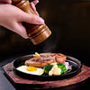 A hand is grinding spices using an adjustable wooden spice mill over a plate of grilled meat and eggs.