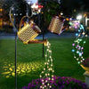 A solar fairy watering can light with glowing wires is displayed in a garden setting.