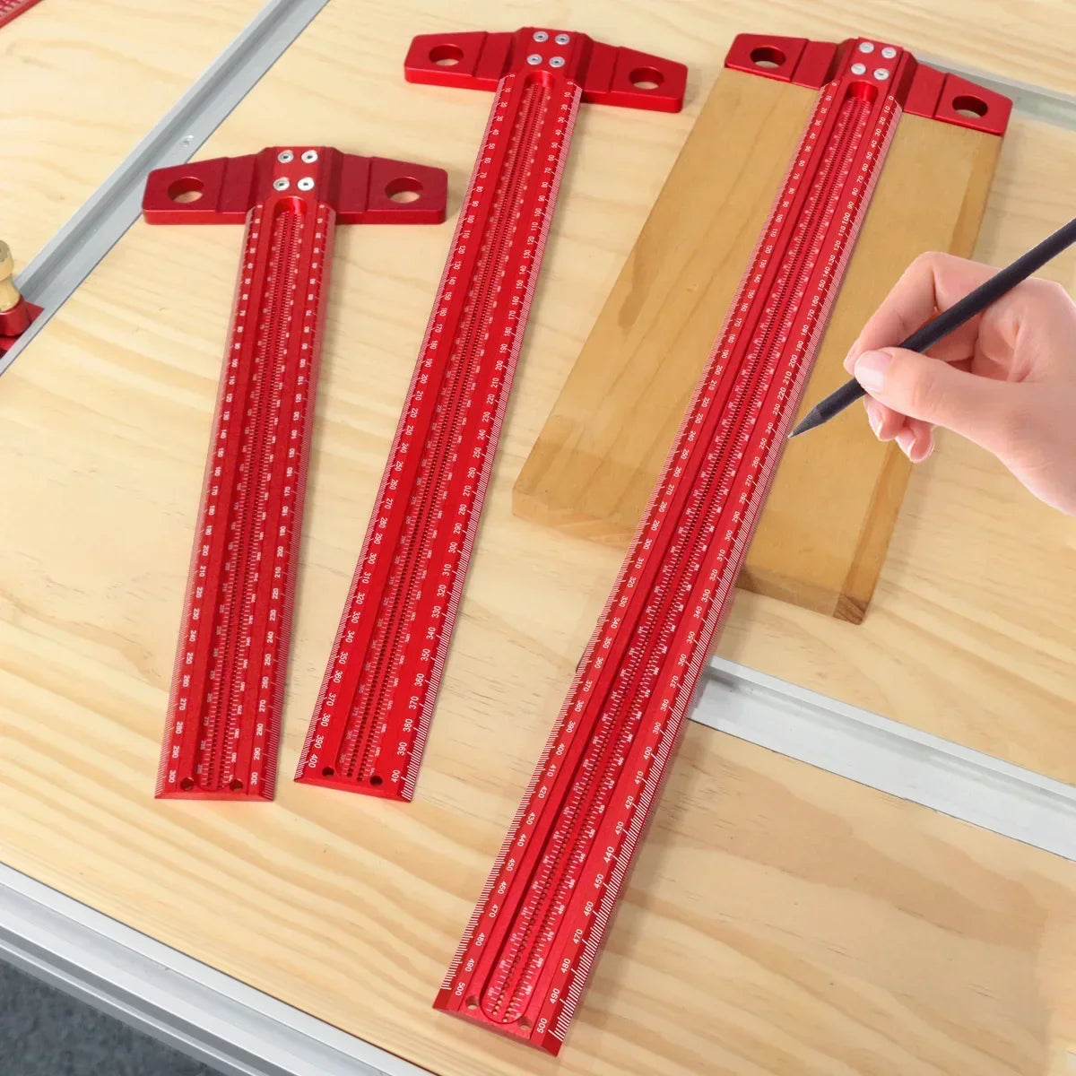 Three red precision marking T-squares are laid out on a wooden surface with a hand holding a black pen.