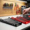 A hand is placing a wrench into a red plastic organizer rack next to various tools on a wooden board.
