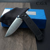 A black folding pocket knife with a silver blade is displayed on a blue box and dark wood surface.