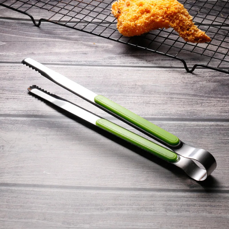 A pair of stainless steel BBQ grill tongs and kitchen clips with green handles resting on a dark wooden surface above a wire 