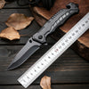 A black multi-functional camping survival knife is displayed next to a ruler on a dark wooden surface with leaves.