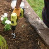 A person is using a yellow and black heavy-duty spiral garden auger drill bit in dark soil next to a concrete edge.