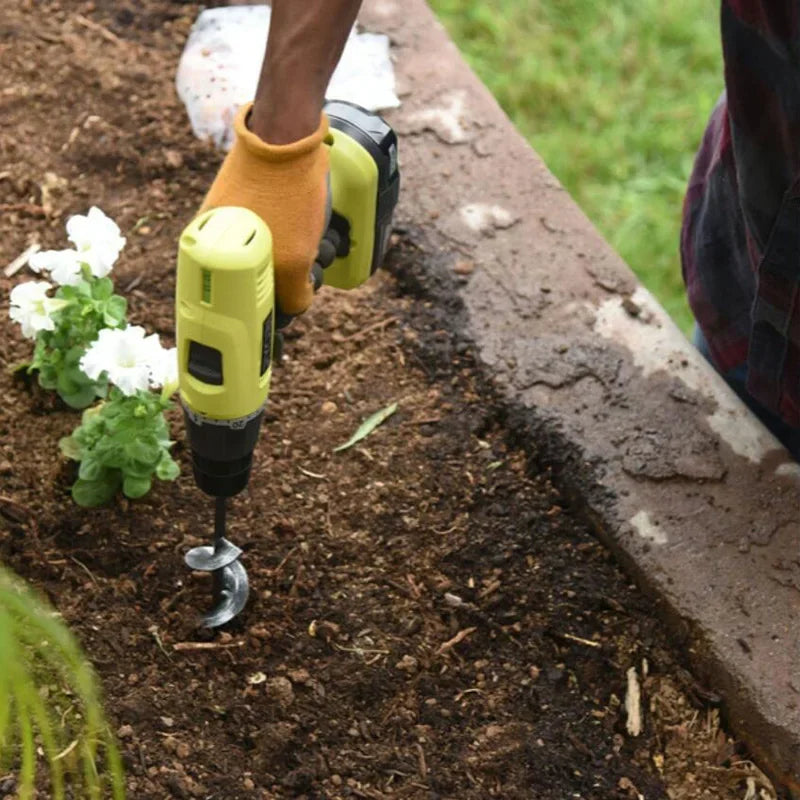 A person is using a yellow and black heavy-duty spiral garden auger drill bit in dark soil next to a concrete edge.