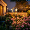 A golden crescent moon solar pathway light illuminates a garden filled with pink and purple flowers at dusk.
