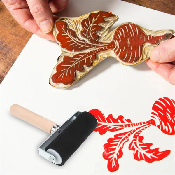 A hand is applying a rubber roller printing tool to transfer a leaf design onto paper.