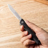A hand holds a black utility folding knife with a silver blade on a light wooden surface.