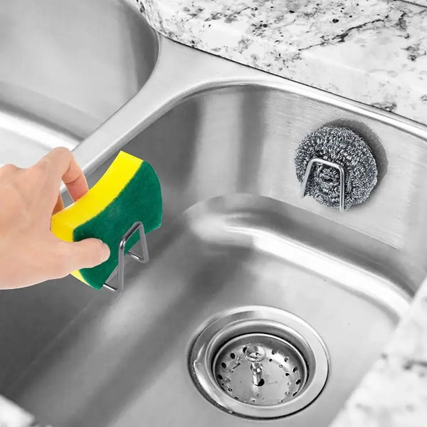 A hand places a yellow and green sponge holder with wall hooks into a stainless steel kitchen sink.